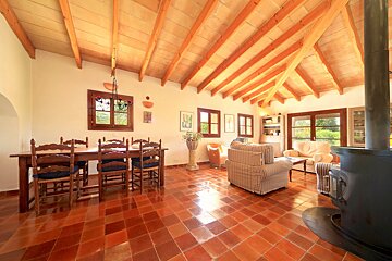A spacious rustic room features a wooden beamed ceiling, terracotta floor, a dining area, a cozy lounge with sofas, and a large wood-burning stove.