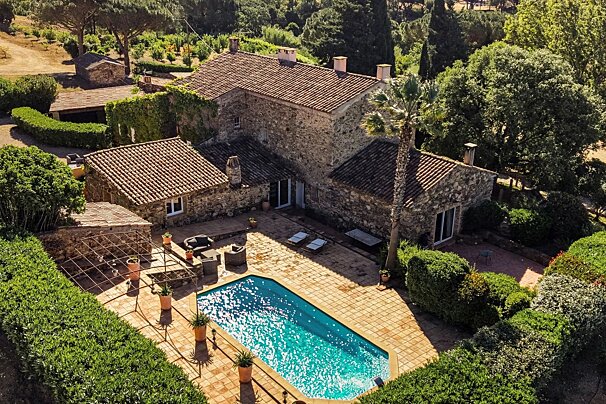 Aerial view of a rustic stone villa with red tile roofs, a sparkling blue swimming pool, and lush green gardens under a sunny sky.