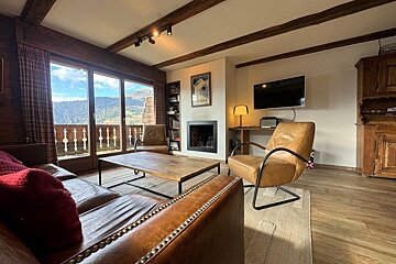 Cozy chalet living room with leather furniture, wooden beams, a fireplace, and large glass doors offering mountain views.