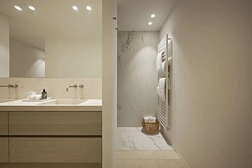 A minimalist modern bathroom with a double sink vanity, a marble shower, and a white towel warmer. Neutral colors and soft recessed lighting.