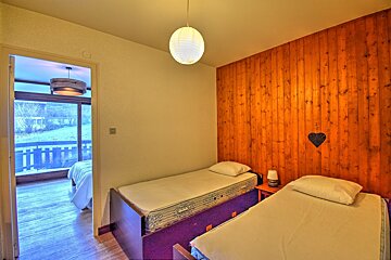 A cozy bedroom with two twin beds, a wood-paneled accent wall adorned with a heart, and a doorway leading to a room with a view of nature.
