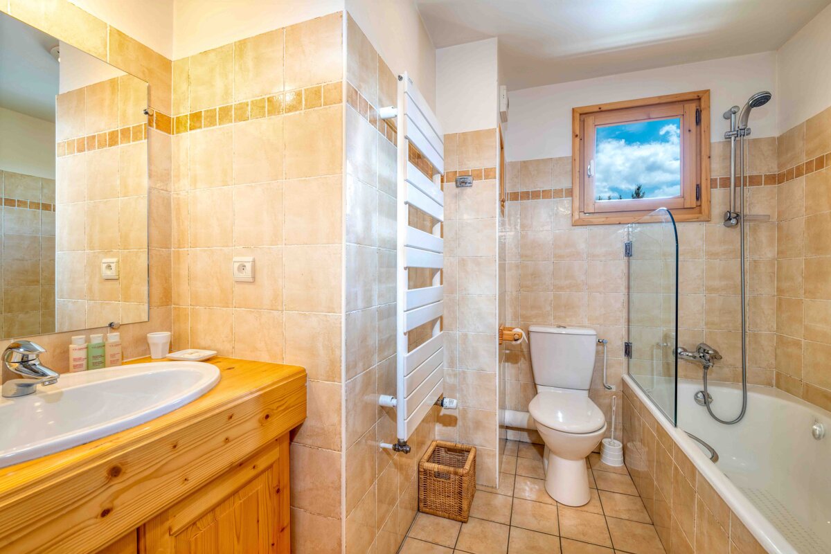 A rustic bathroom with beige tiles, wooden vanity, mirror, toilet, and bathtub with shower. A window shows a cloudy sky.
