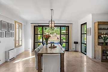A bright dining room features a long wooden table, white chairs, and expansive glass doors opening to a lush garden. Light tiled floors and white walls create an airy feel.