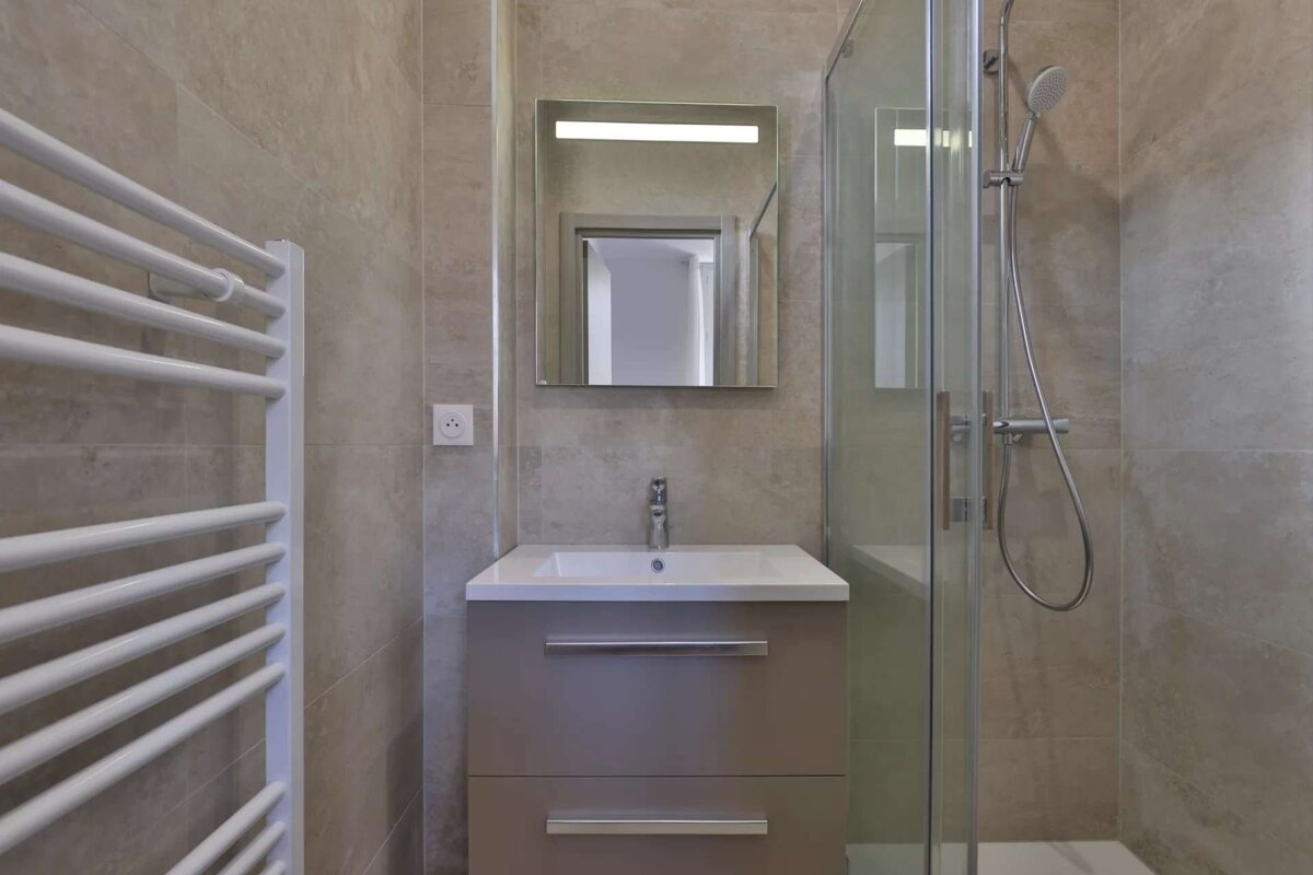 A modern bathroom with beige tiled walls, a white towel warmer, a vanity with sink, a lighted mirror, and a glass-enclosed shower.