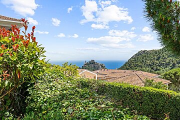 Lush green garden overlooks tiled roofs, a distant hilltop castle, and a calm blue sea under a partly cloudy sky.