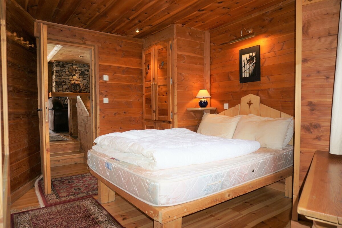 Cozy wooden cabin bedroom featuring a double bed with white bedding, wood-paneled walls and ceiling, and an open doorway.