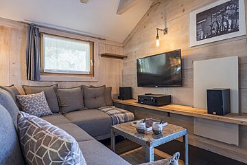 A warm, rustic living room with a large gray sectional sofa, TV, and industrial-style coffee table. Light wooden paneled walls and a window create a cozy ambiance.