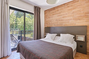 Modern bedroom with a bed, wooden wall, and a balcony view of green trees.
