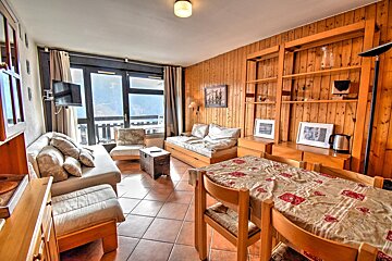 Cozy chalet-style room featuring wood-paneled walls, a dining table, tiled floors, multiple sofas, and large windows with scenic views.