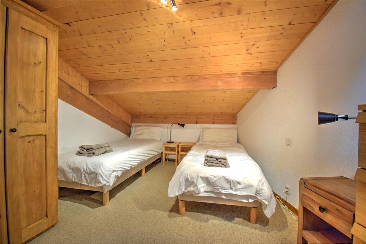 A cozy attic bedroom with two single beds, white bedding, light wood paneling on the ceiling and walls, and light brown carpet.