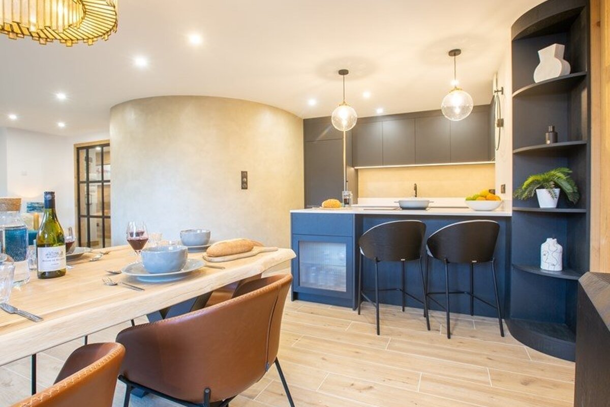 A chic modern kitchen and dining room featuring a wooden table, leather chairs, and a dark blue island with globe pendant lights.
