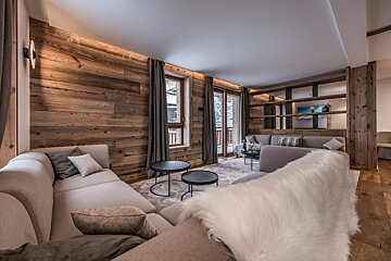 A stylish, modern chalet living room with warm wood walls, plush light sofas, large windows to a snowy view, and an open shelving unit.