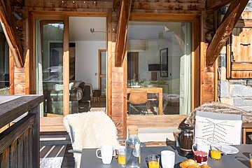 A sunny balcony table set for breakfast outside a cozy, modern wooden chalet with large glass sliding doors.