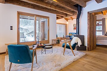 Cozy chalet living room featuring teal armchairs on a plush rug, a wood-burning stove, and glass doors overlooking a snowy winter scene.