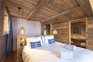 Chalet-style bedroom featuring rich wood walls/ceiling, a comfortable bed with plaid pillows, modern lighting, and a visible en-suite bathroom.