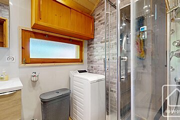 A compact bathroom featuring a glass shower, washing machine, wooden window, cabinet, and a stone-look tiled accent wall.