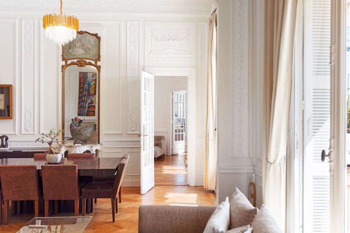 An elegant, sunlit room with ornate white paneled walls, a large mirror, and a gold chandelier. A dining table sits on a beautiful herringbone floor.