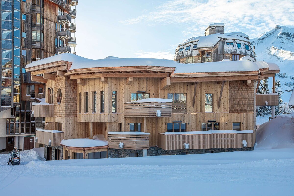 Snowy modern wooden chalets and ski resorts, with a snow-capped mountain range under a blue sky.