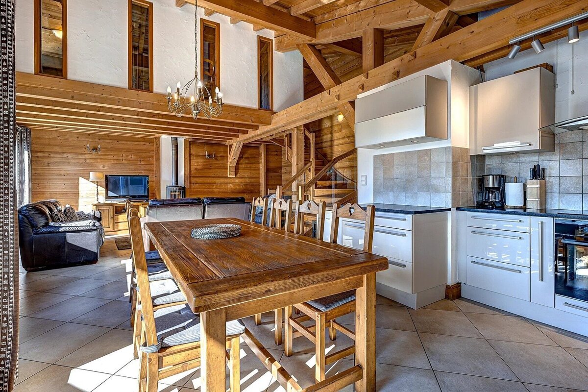Spacious, open-plan chalet interior with rustic wooden walls and beams, a large dining table, living area, and modern white kitchen.