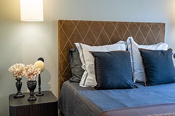 Cozy bedroom with a brown quilted headboard, blue and white pillows, and a nightstand adorned with coral under a wall lamp.