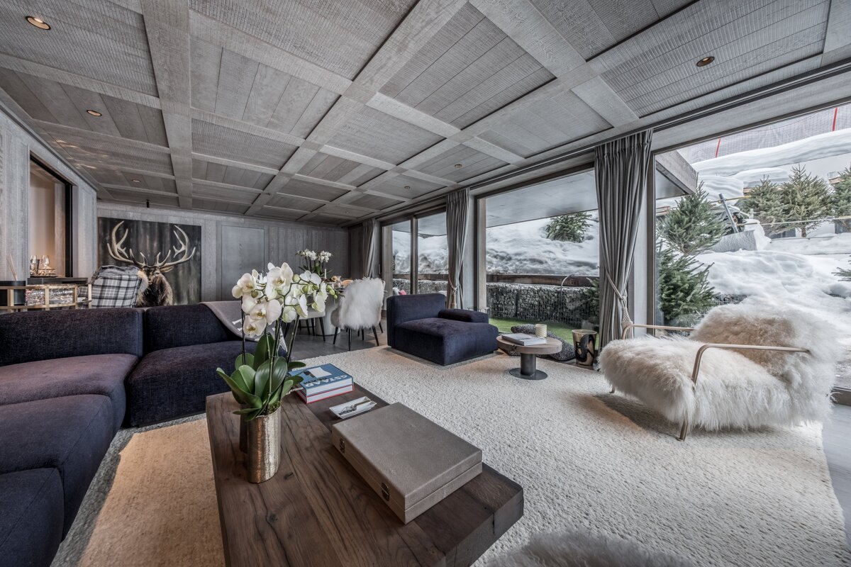 Cozy luxury chalet living room with a wooden ceiling, plush seating, and large windows overlooking a snowy landscape.