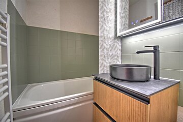 Modern bathroom with a white bathtub, sage green tiled walls, a wood vanity with a black vessel sink, and an illuminated mirror.