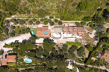 Drone shot of a sprawling, multi-building estate with terracotta roofs, two pools, terraced gardens, and lush green hillsides.