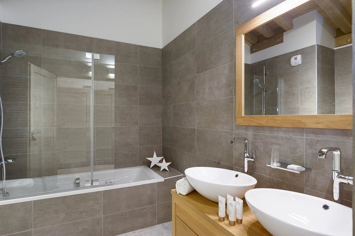 Modern bathroom with gray tiles, a bathtub with a glass screen, and a wooden vanity featuring two white vessel sinks and a mirror.