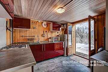 A cozy kitchen with wood paneling and red cabinets, opening to an open door with a stunning snowy mountain and forest view.