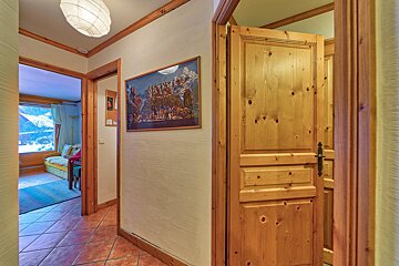 A cozy hallway with warm wooden doors/trim, terracotta tiled floor, and a framed mountain print. A room with a window showing a snowy view is visible.