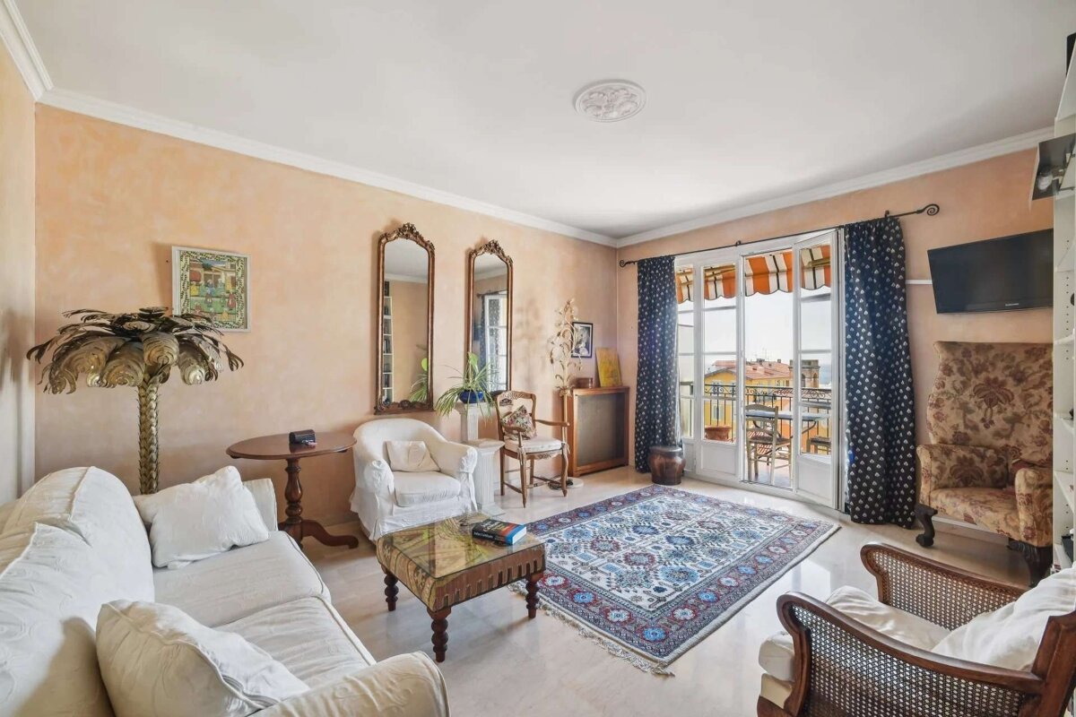 A warm, eclectic living room with peach walls, white & patterned furniture, ornate mirrors, a colorful rug, and French doors to a balcony.