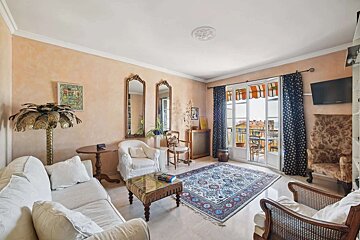 A warm, eclectic living room with peach walls, white & patterned furniture, ornate mirrors, a colorful rug, and French doors to a balcony.