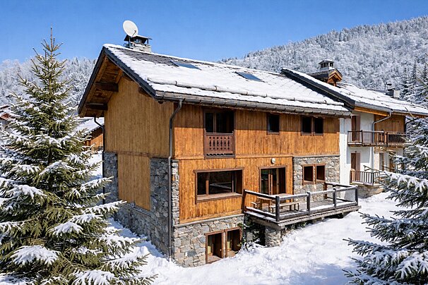 Snow-covered wooden alpine chalet with a stone base, surrounded by snowy pine trees under a bright blue sky.