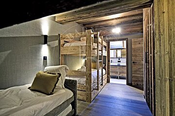 A rustic cabin-style bedroom featuring wooden bunk beds, a single bed in the foreground, and an en-suite bathroom visible down the hall.