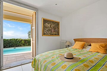 A bright bedroom with a straw hat on a patterned bed, opening onto a sunny patio with a sparkling swimming pool.