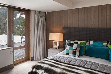 Cozy bedroom with wooden wall, colorful pillows, and a glass door to a snowy deck and forested mountain view.