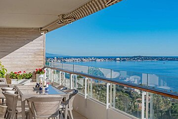 Panoramic view from a sunny balcony with outdoor dining, showing a vast blue sea, distant city, and coastline.