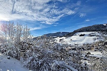 A picturesque winter scene shows snow-covered mountains, a village, and frosty trees under a bright blue sky with scattered clouds.