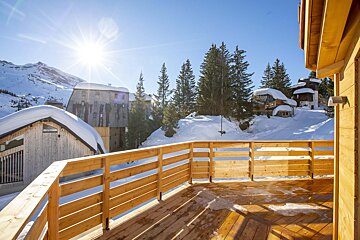 Sunny winter view of snow-covered chalets and pine trees from a wooden balcony in a mountain resort.
