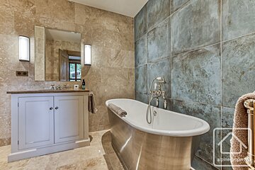 A modern rustic bathroom featuring a freestanding silver bathtub, a light vanity, and textured beige and distressed blue-grey wall tiles.