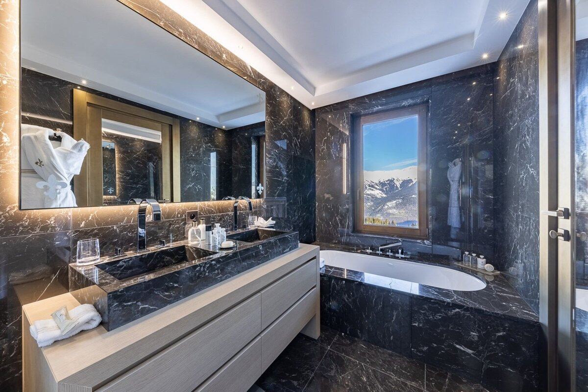 A luxurious black marble bathroom features a double vanity, sleek bathtub, and a large window showcasing stunning snowy mountain views.