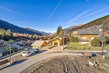 Sunny mountain village with wooden chalets and winding roads, surrounded by forested hills under a bright blue sky with airplane contrails.