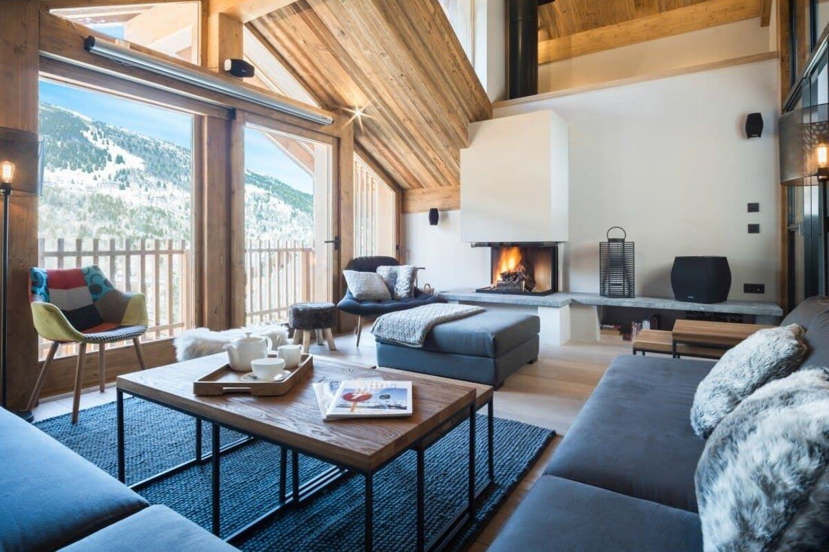 A warm, modern chalet living room with a fireplace, comfy sofas, and expansive windows overlooking snowy mountains.