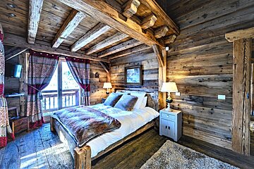 A cozy, rustic wooden bedroom featuring exposed beams, a large bed with a fur throw, bedside lamps, and a balcony with red patterned curtains.