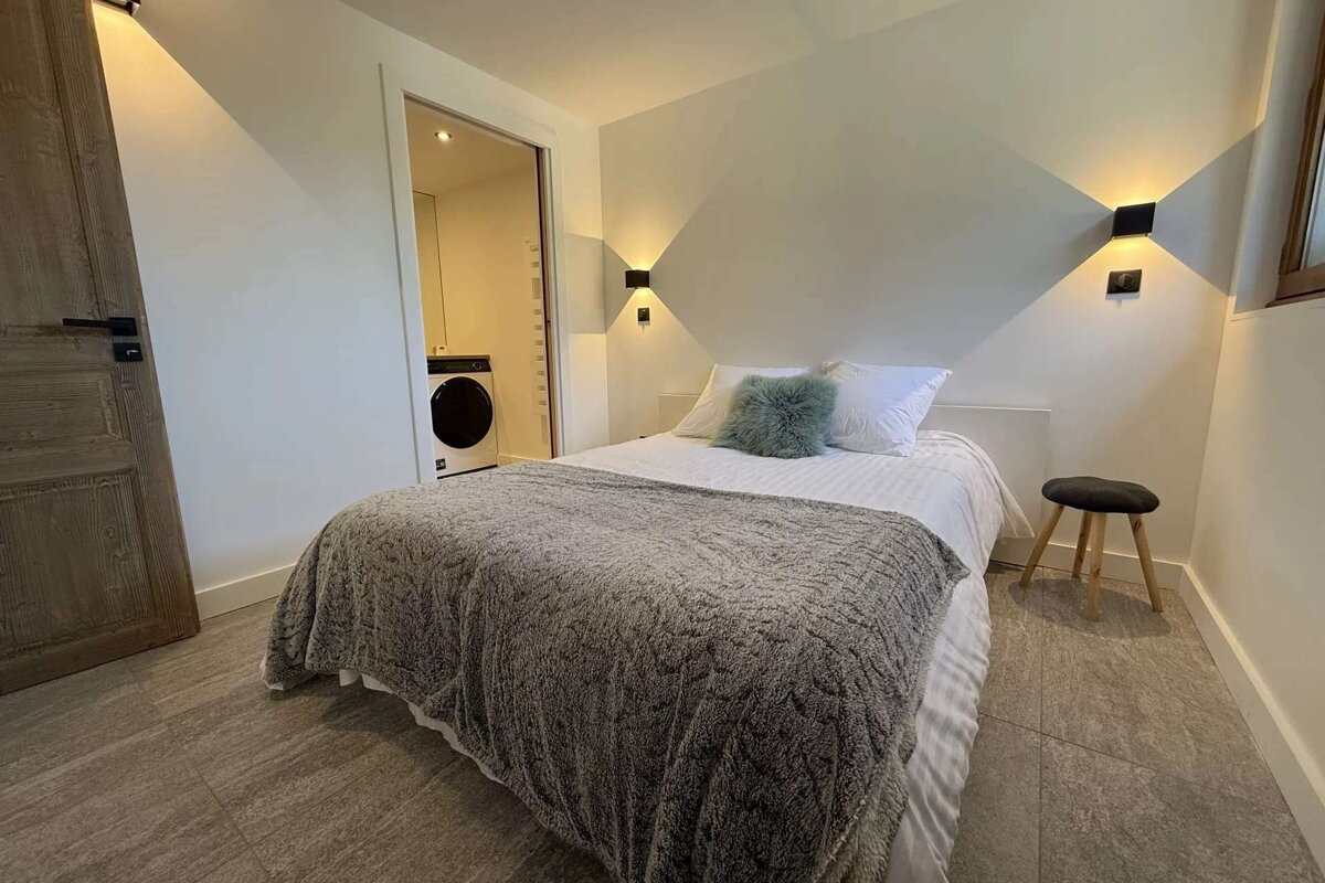 A cozy bedroom features a bed with white sheets and a grey fuzzy blanket. Wall sconces illuminate the space, and a washing machine is visible through an open doorway.