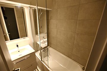 A modern, tiled bathroom with a white bathtub and glass shower screen, a vanity with a sink, and a mirror.