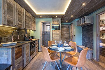 A cozy rustic apartment featuring a wooden kitchen, warm overhead lighting, and a round dining table set for four with tan chairs.