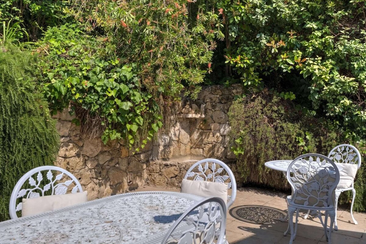 Sunny outdoor patio with ornate white metal tables and chairs, nestled against a verdant, stone wall covered in plants.
