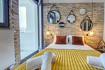 Cozy bedroom with a yellow bed, orange cushions, and a textured wall adorned with round mirrors and hanging lights. A balcony view is visible.