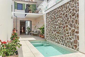 A sunlit modern courtyard with a plunge pool, a textured stone wall, outdoor seating, potted plants, and a building with a balcony.